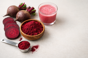 Editorial hero photo of beetroot powder in a bowl with fresh beets, a measuring spoon, and a pink beverage on a neutral background.